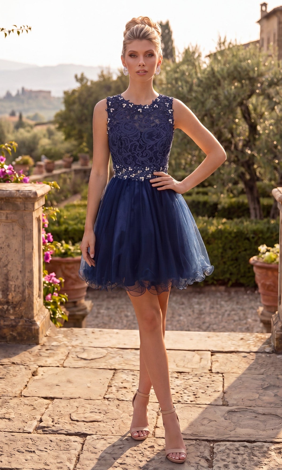 A woman stands outdoors on a stone path, wearing the Short Homecoming Dress: Chicas C9350 in navy. The dress features a sleeveless illusion lace bodice adorned with floral details and an elegant a-line tulle skirt, paired with nude heels. Lush greenery and distant hills create a scenic backdrop.